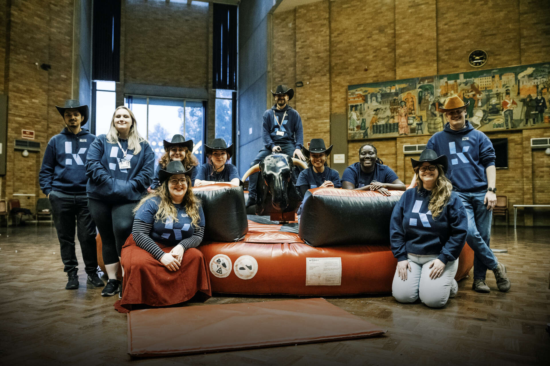 University of Kent ResLife team wearing cowboy hats gathered around a mechanical bull inside a campus hall.