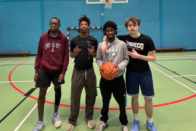 Four basketball players pose together on an indoor court, one holding a ball, beneath a hoop against a blue wall.