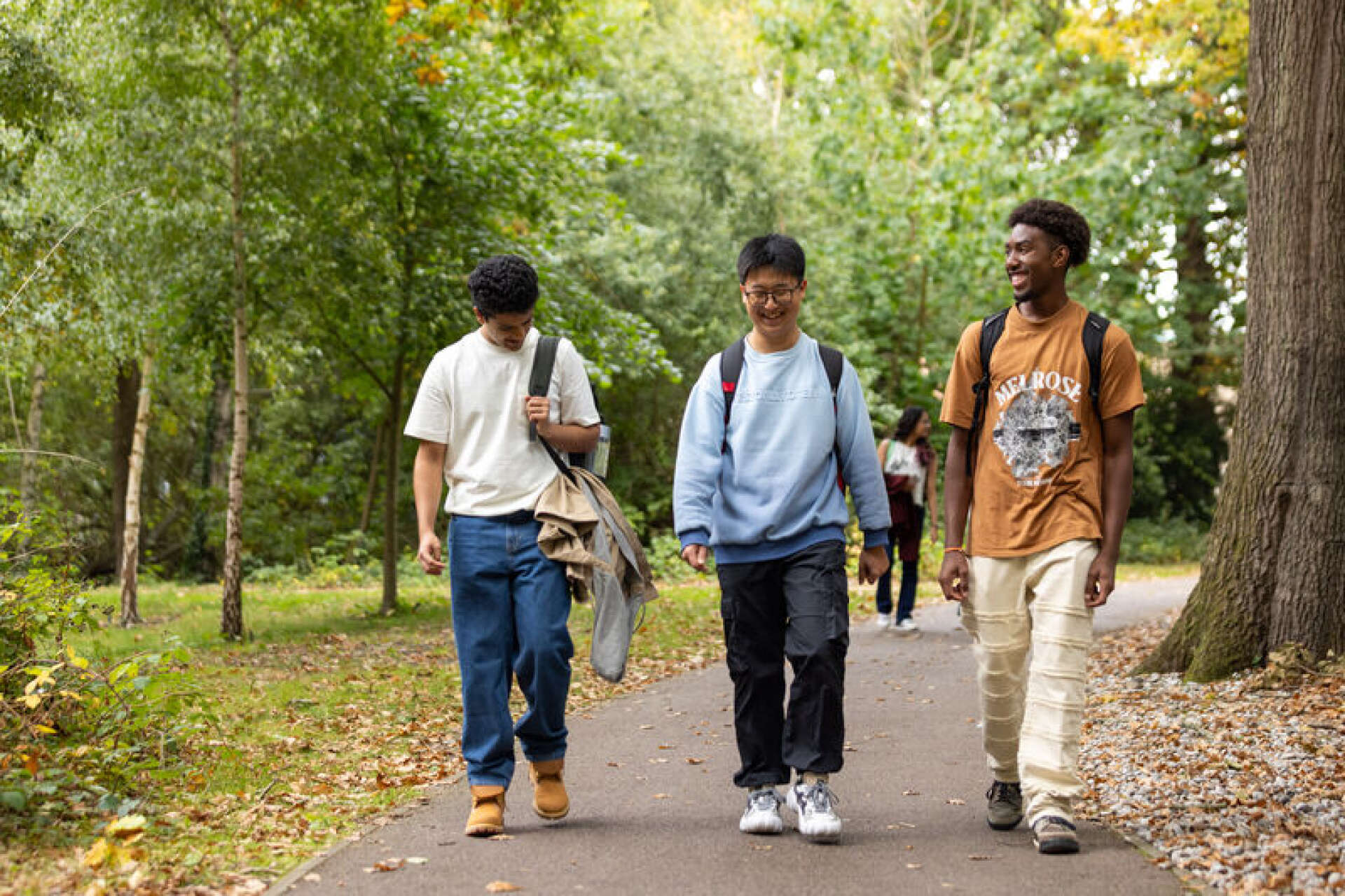 Gemini said Three young men with backpacks walk and laugh along a tree-lined paved path in a park.