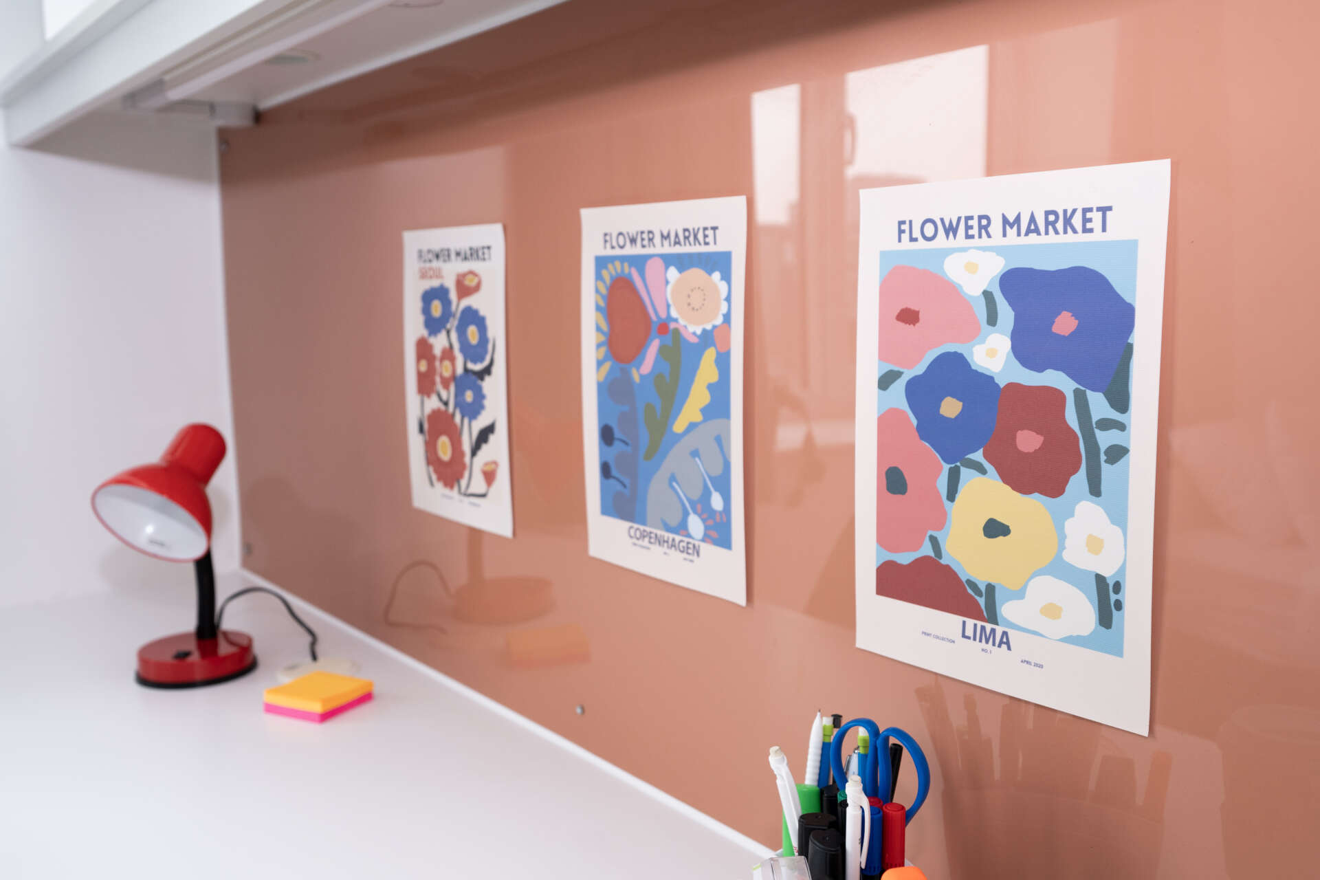 A white desk with a red lamp and office supplies sits before a peach backsplash featuring three "Flower Market" art prints.