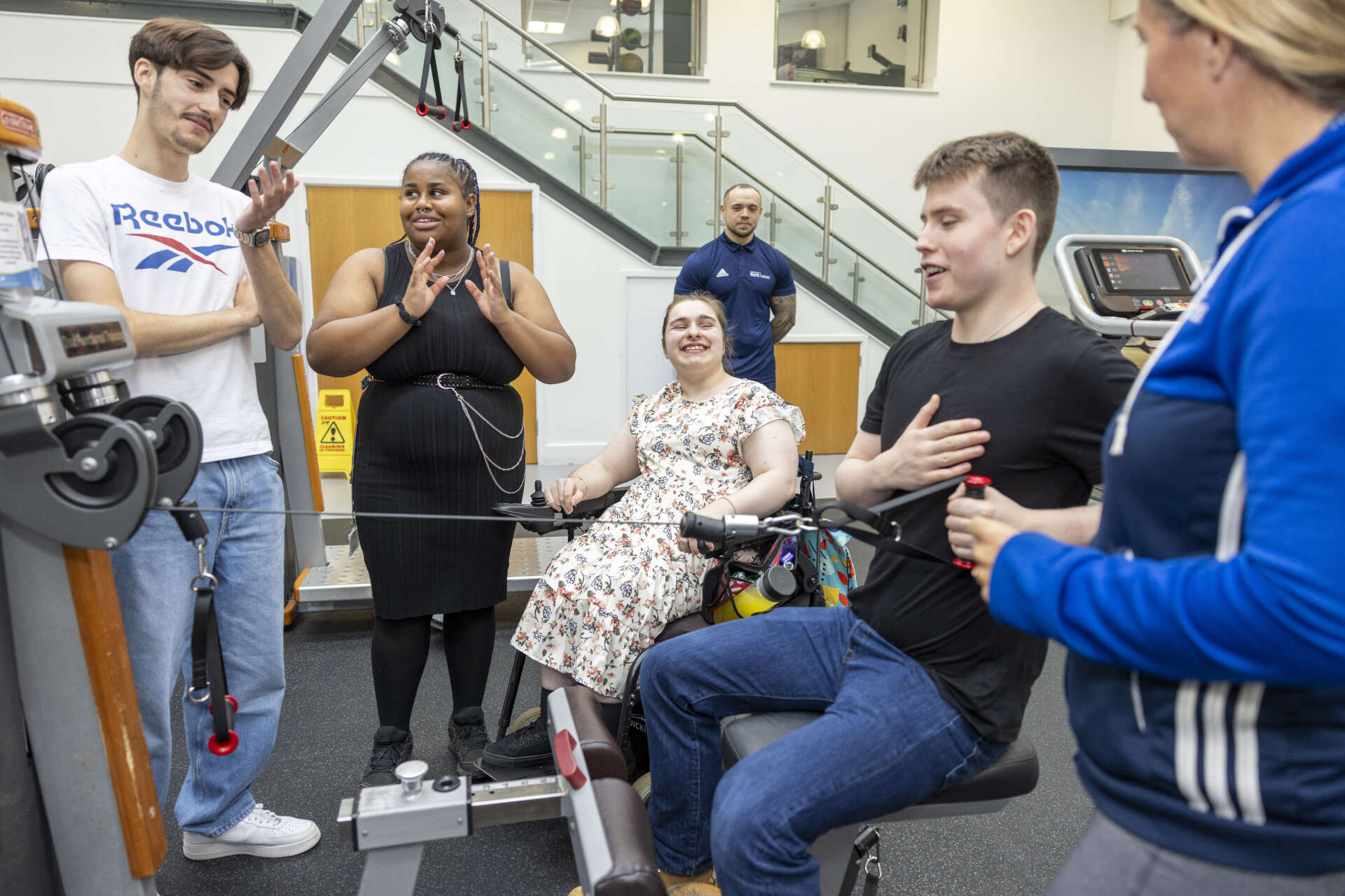 Wheelchair user smiling while watching a young person use a weights machine with staff overseeing