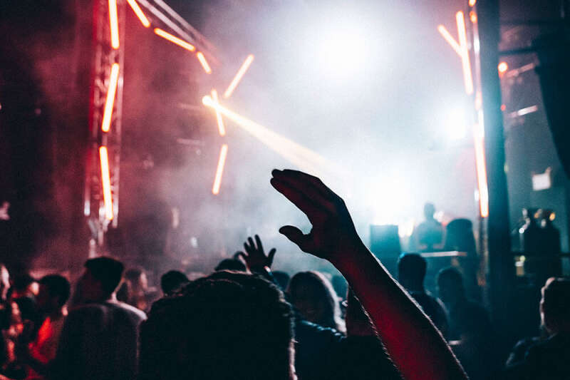 A dance party at the campus nightclub, a young man raises his hand in the air in front of the decks.