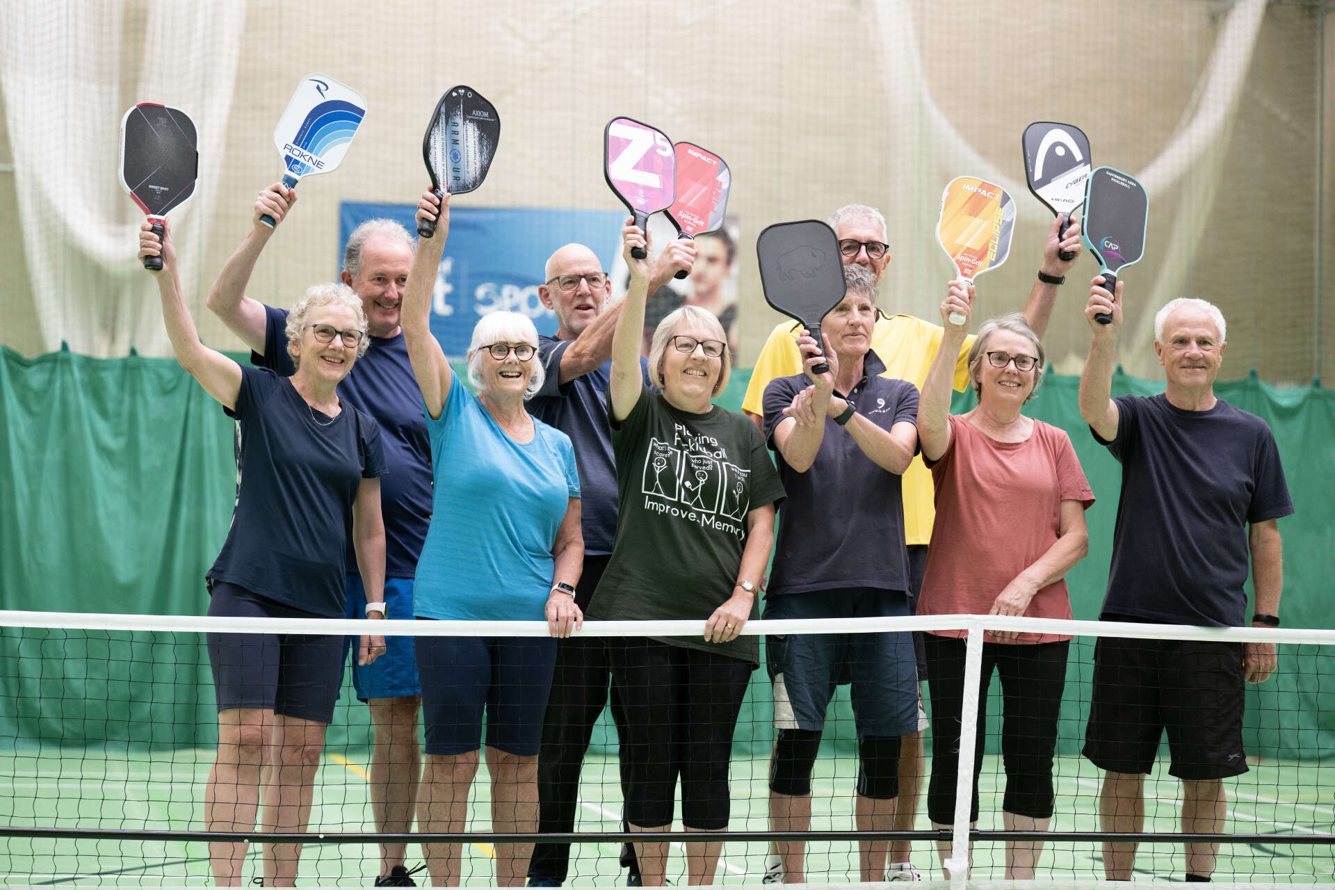Nine pickleball players standing on the court behind the net holding rackets in the air and smiling