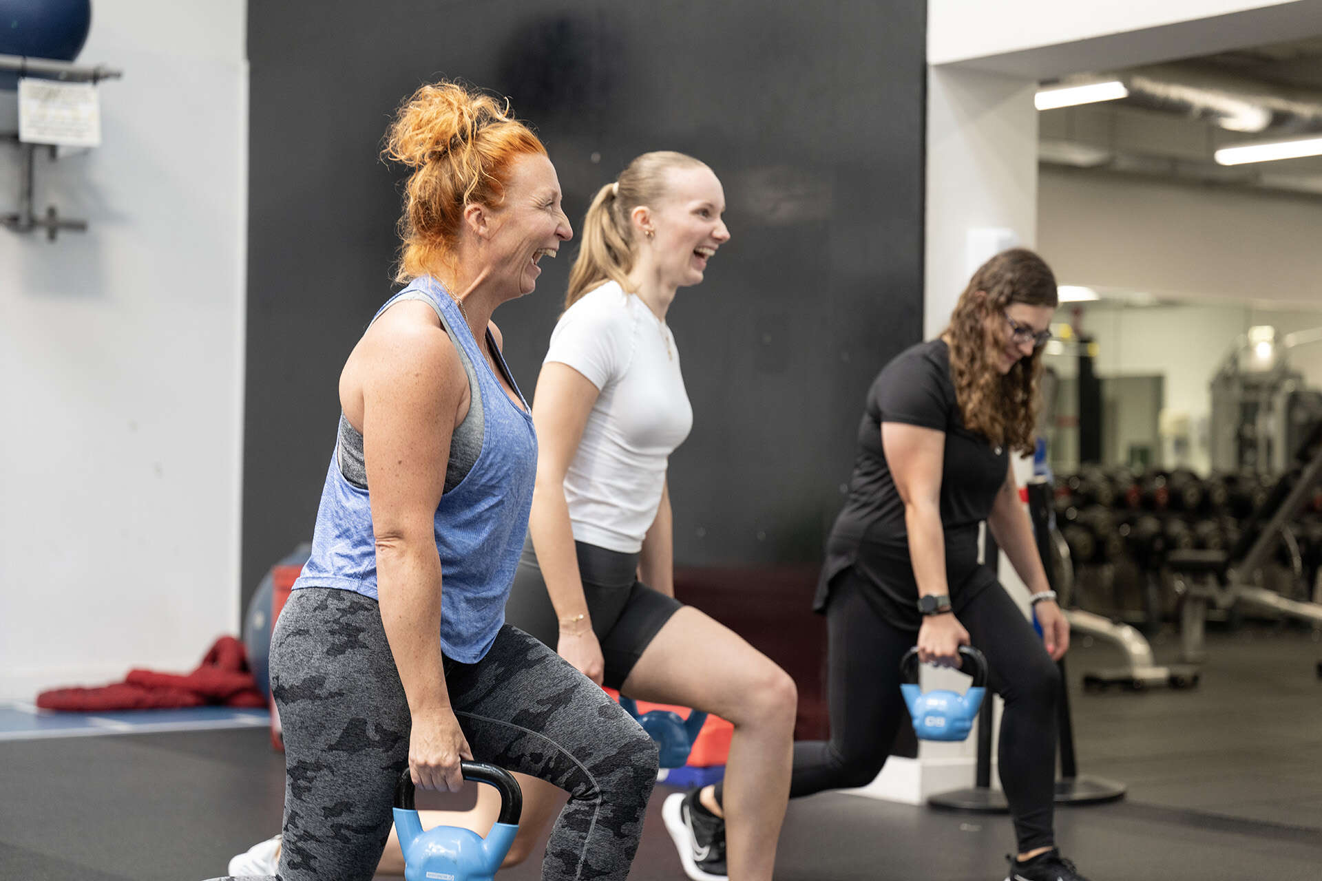 Two people lunging while holding kettlebells and smiling, looking away from the camera
