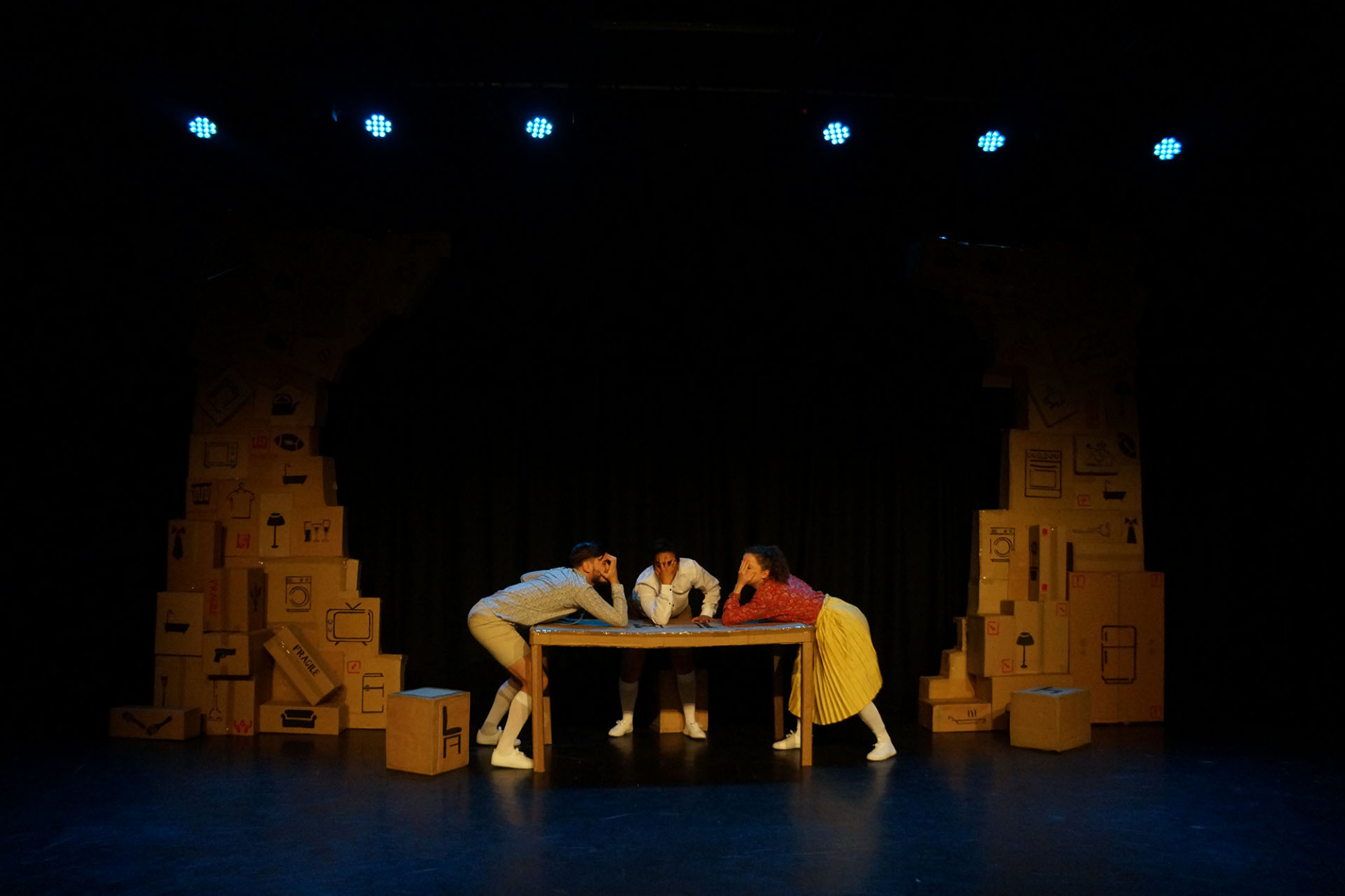 Three performers lean over a table with cardboard boxes stacked on each side of the stage