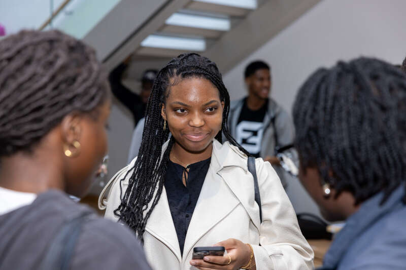 A woman holding her phone and listening to someone who is speaking to her, whilst someone else is also present.