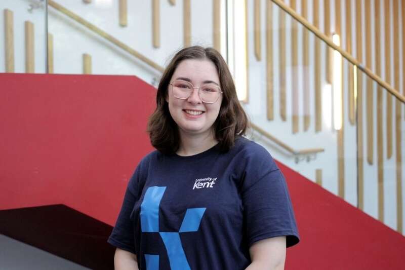 Jess in a blue Kent branded t-shirt, posing by the stairs in our Kennedy building.