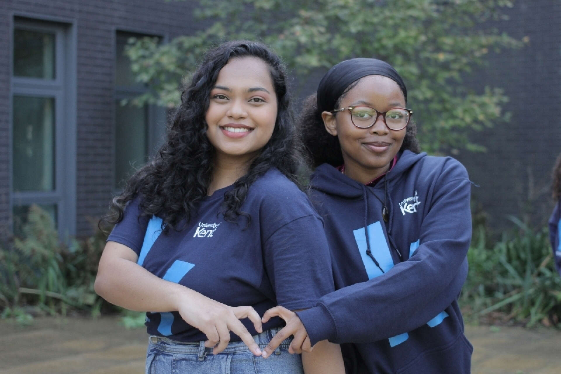 ResLife Ambassadors Mishti and Panashe posing in branded tshirt and hoodie in front of our Kennedy building.