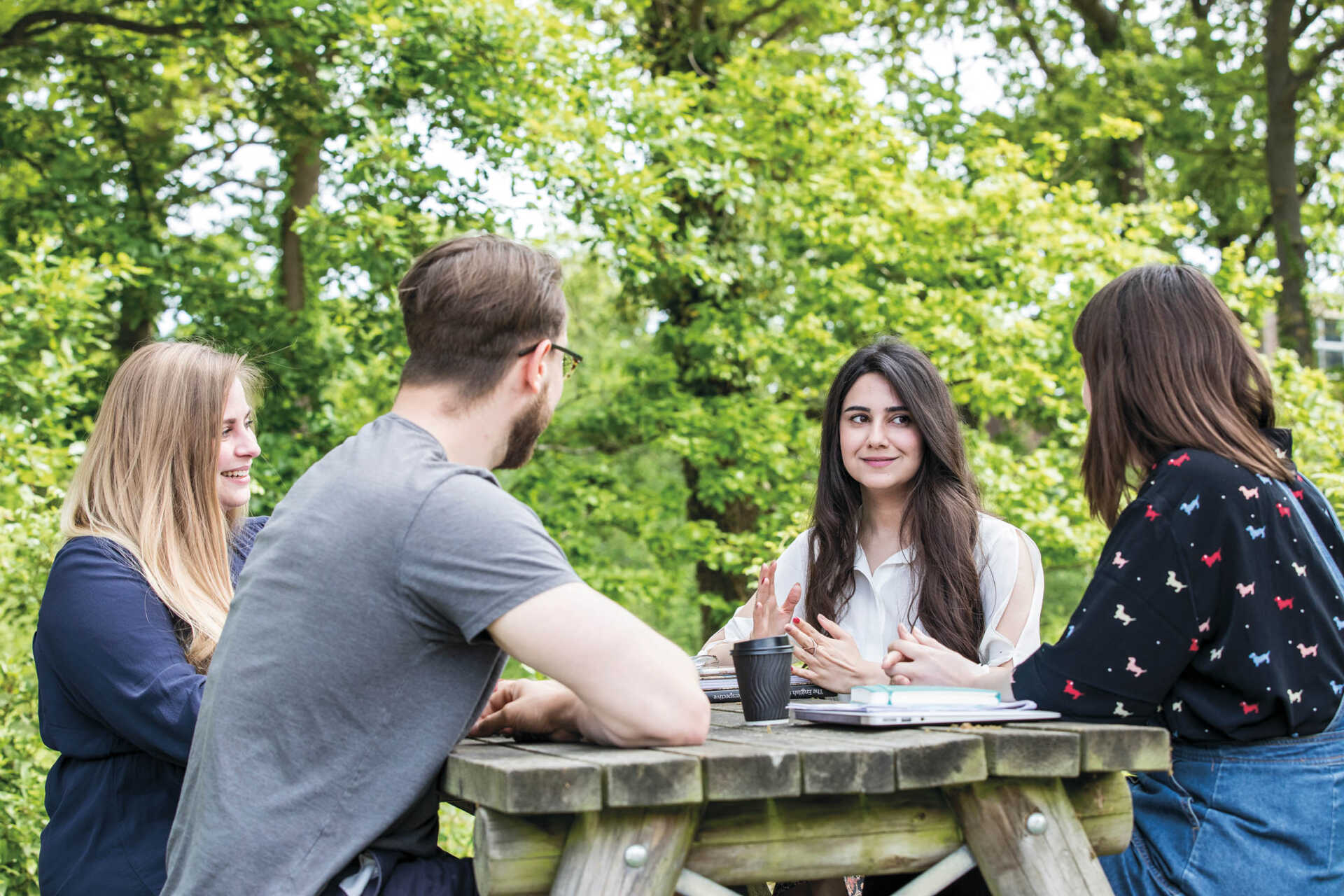 Four students sit around a table outside. The are surrounded by greenery and smile towards each other.