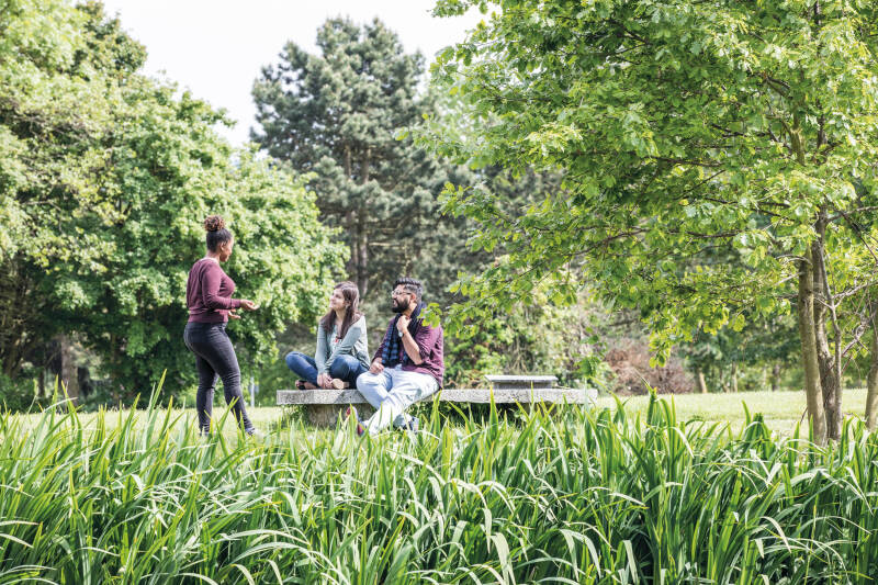 A group of 3 students sat outside on a bench talking surrounded by trees.
