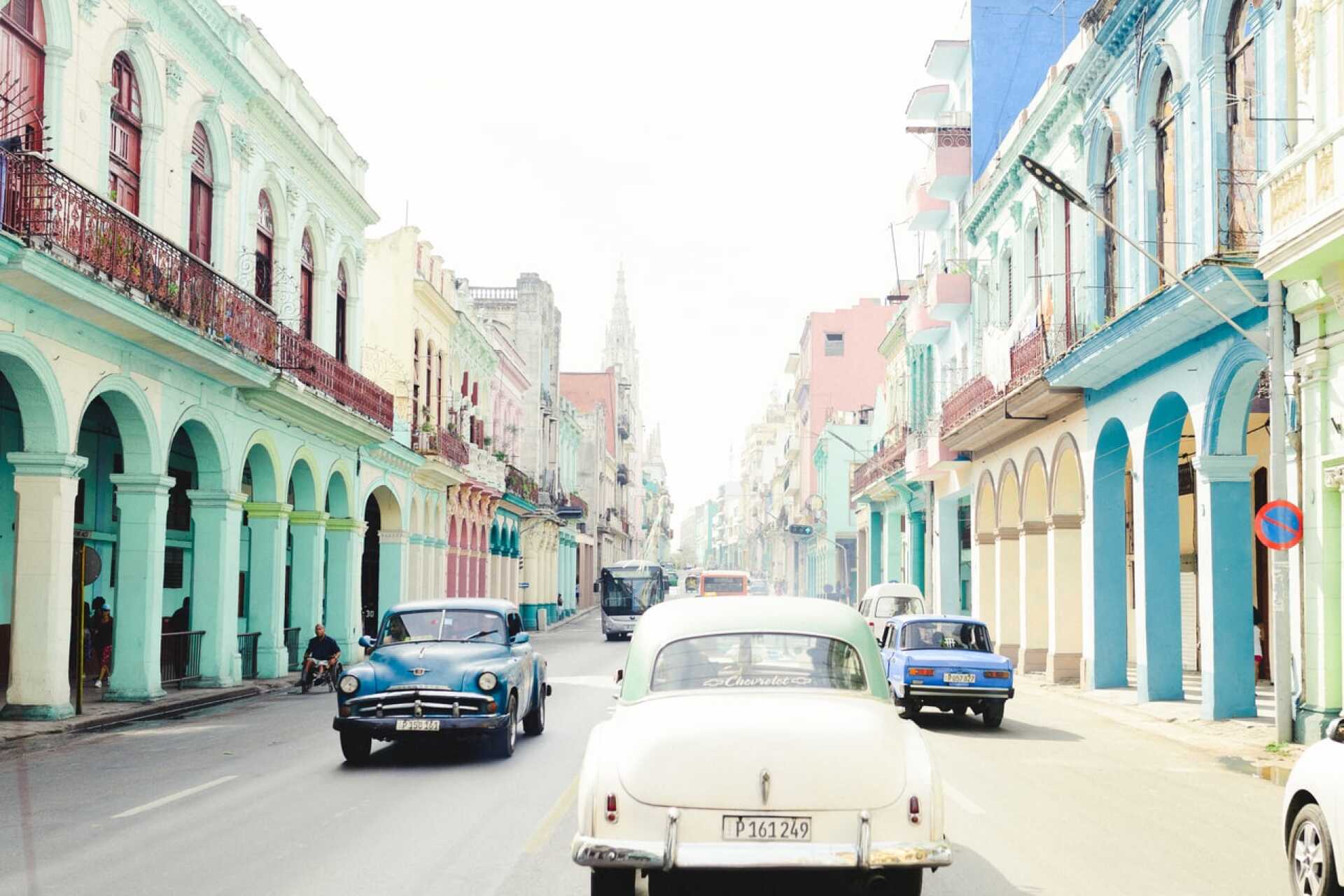 Cars drive along a street of brightly-coloured houses in Cuba