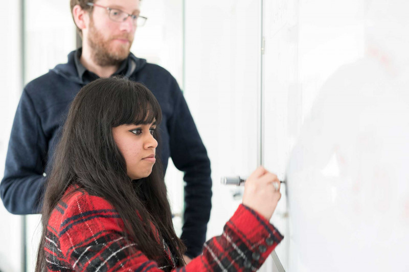 Girl with long dark hair and red check jacket writing on whiteboard