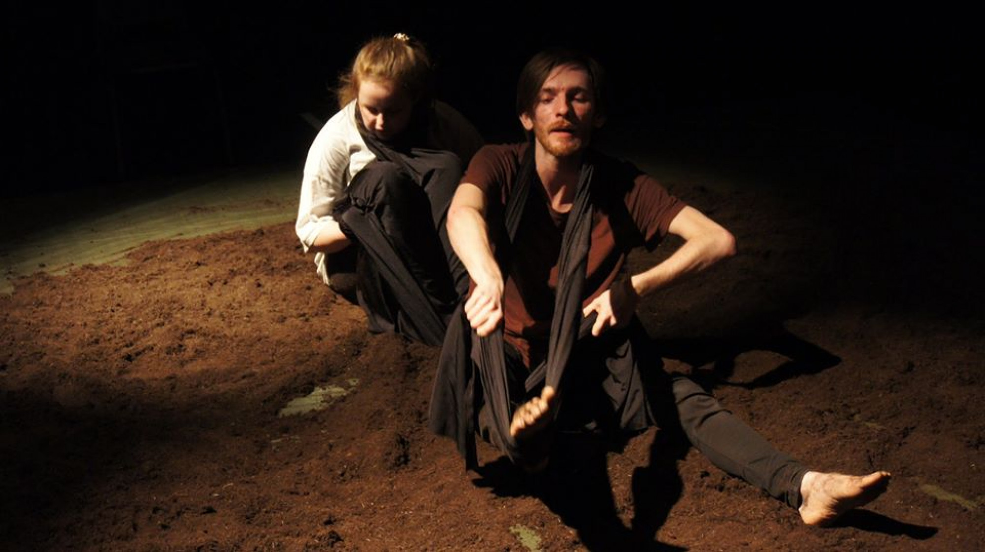 Two performers sit in moody lighting surrounded by black drapes and mud.