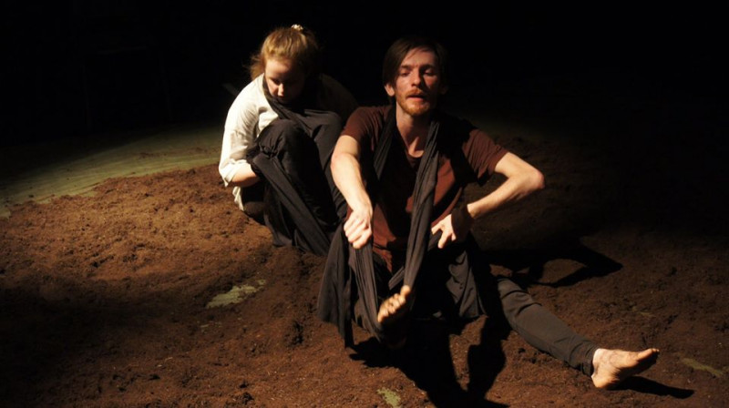 Two performers sit in moody lighting surrounded by black drapes and mud.