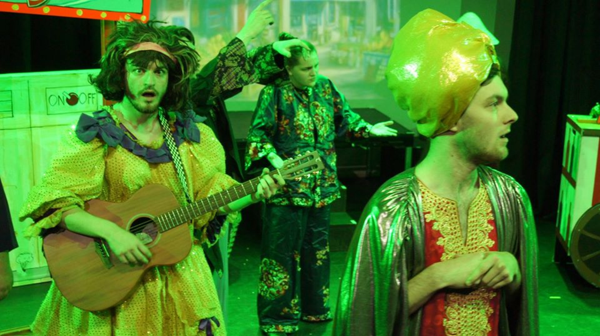 The panto dame plays a guitar, while the villain looks offstage.