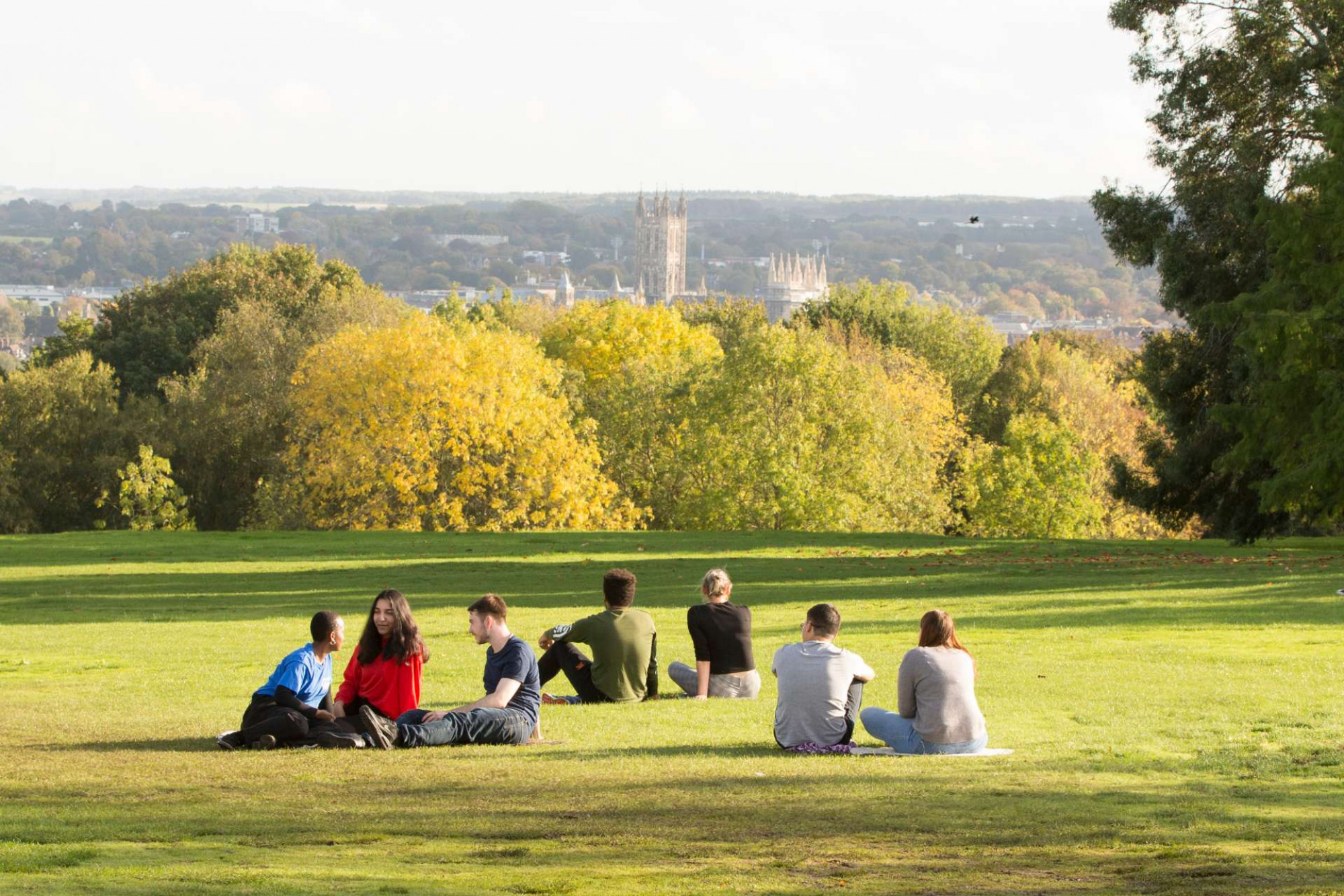 Students sitting on grass looking at beautiful view from the Canterbury campus across the city to the Cathedral in autumn