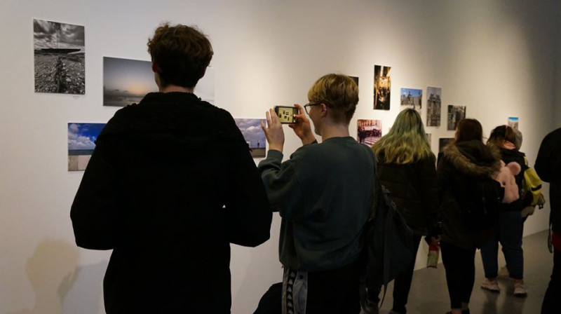 Students stand facing away from the camera, looking at a white wall with photos on. One is taking a photo with a smartphone.