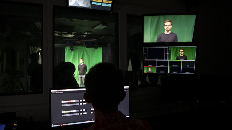 In the editing suite. The room is dark, several screens are visible showing a person standing in front of a green screen.