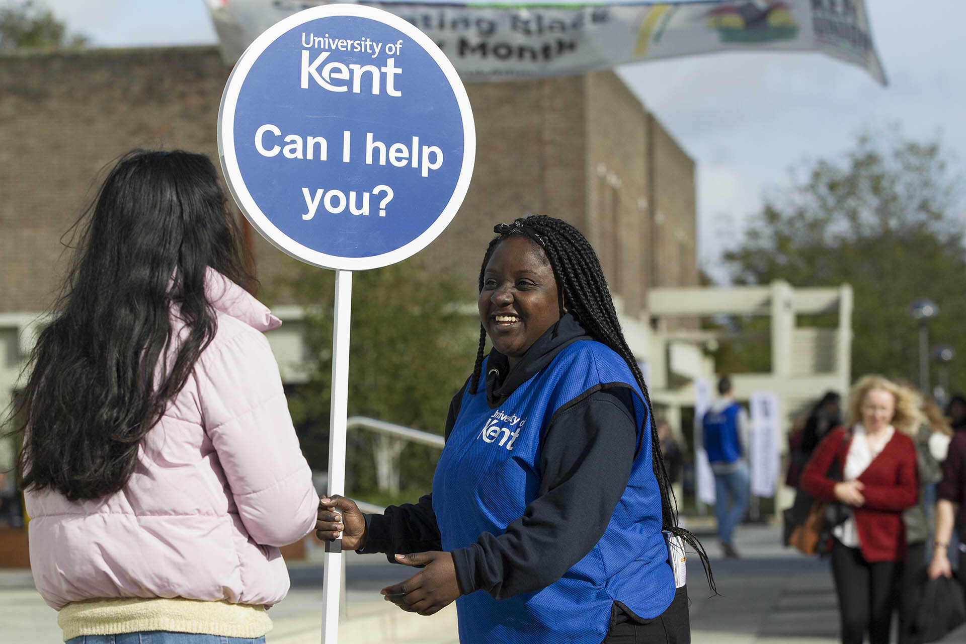 A smiling student ambassador holding a Can I help you? lollipop sign